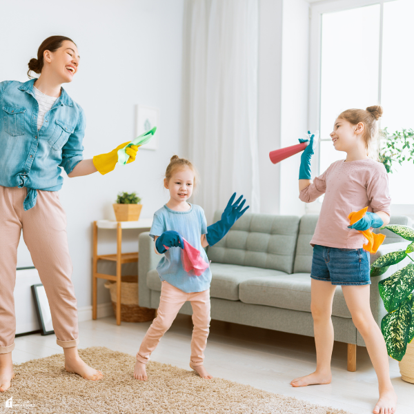 A mother and her two daughters wearing rubber gloves playfully clean their living room with spray bottles and cloths.