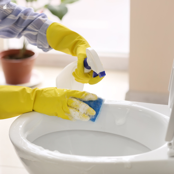 A person wearing yellow gloves scrubbing a white toilet with a blue sponge while spraying cleaner during a bathroom deep clean.
