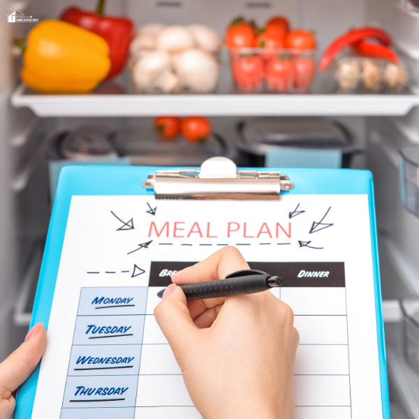 A hand writes a weekly meal plan on a clipboard placed in front of an open refrigerator filled with fresh produce.
