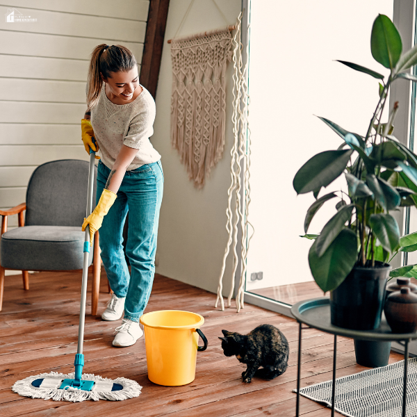 A woman wearing gloves mops a wooden floor near a window while a curious cat watches, highlighting indoor cleaning during seasonal chores.