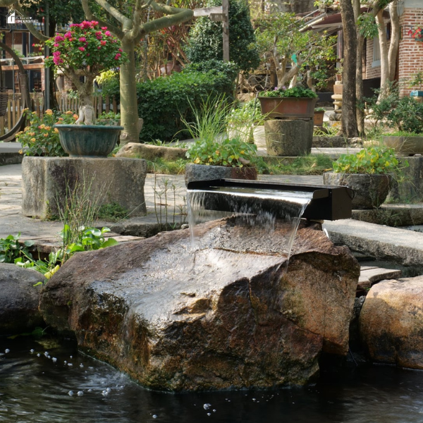 A black waterfall bucket sits on a large rock, cascading water into a backyard pond surrounded by natural stones.