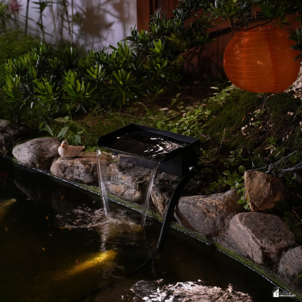 A lit backyard pond features a black waterfall bucket flowing gently over rocks, with koi fish visible beneath the surface.