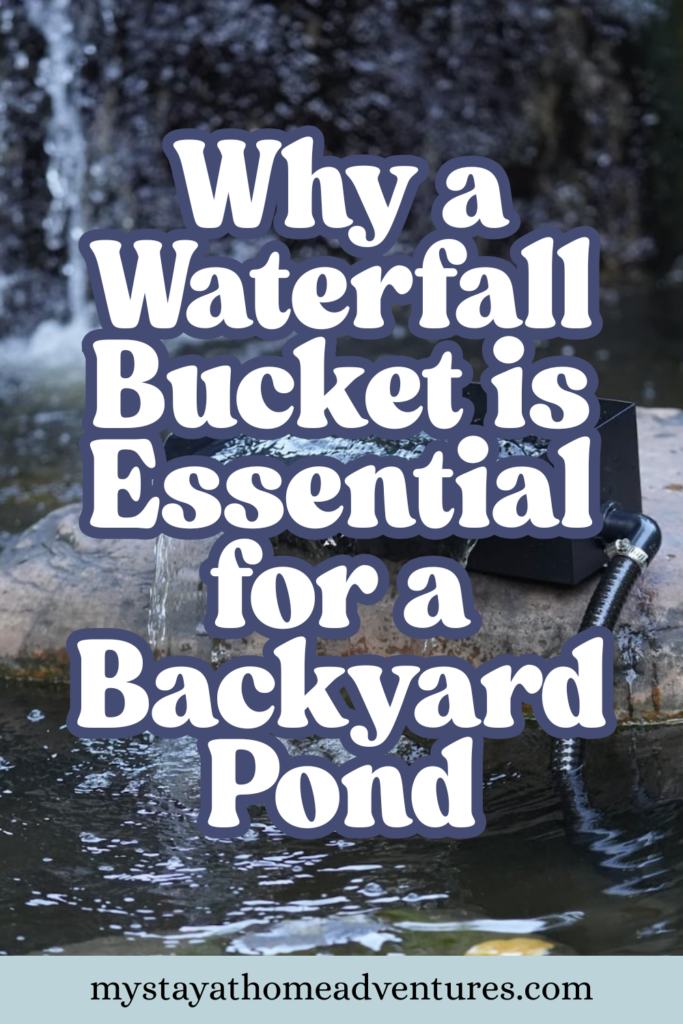 A waterfall bucket flows over a large rock into a garden pond, surrounded by lush plants and stone landscaping in a rustic yard.