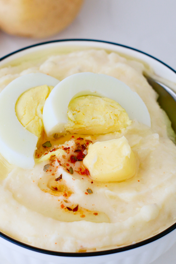 Bowl of Puerto Rican mashed potatoes with eggs, topped with sliced hard-boiled egg, a pat of butter, and adobo seasoning—an easy Latin mashed potato side dish perfect for comfort food lovers and holiday meals.