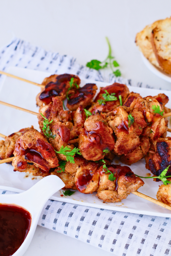 Puerto Rican chicken pinchos topped with BBQ sauce and fresh herbs, served on skewers with a side of bread and dipping sauce on a white platter.