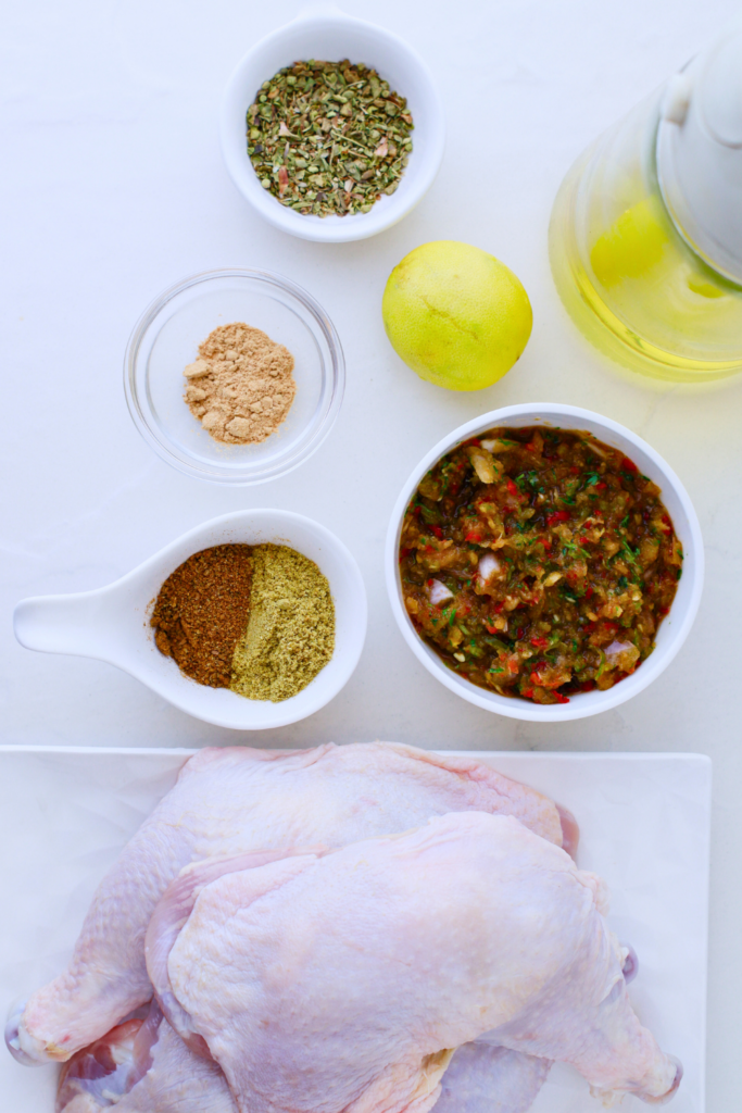 Raw chicken leg quarters, sofrito, olive oil, lime, and traditional Puerto Rican spices including oregano, cumin, coriander, and garlic powder arranged on a white surface.
