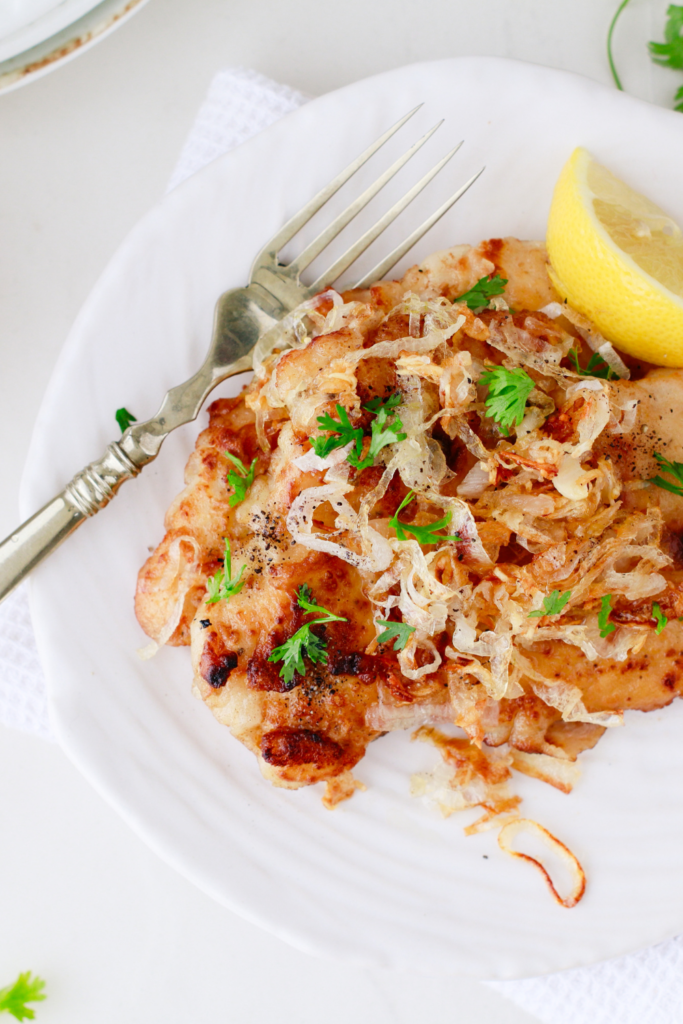 A serving of Cuban lemon chicken topped with caramelized onions and fresh parsley, plated with a lemon wedge and vintage fork.