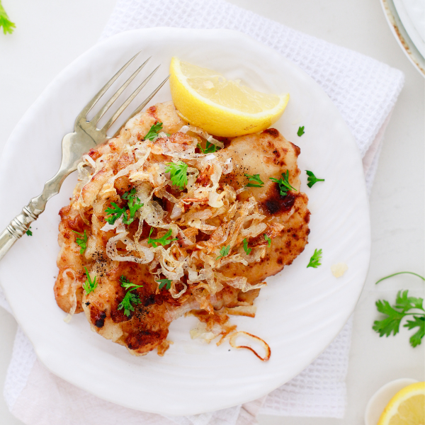 A serving of Cuban lemon chicken topped with caramelized onions and fresh parsley, plated with a lemon wedge and vintage fork.