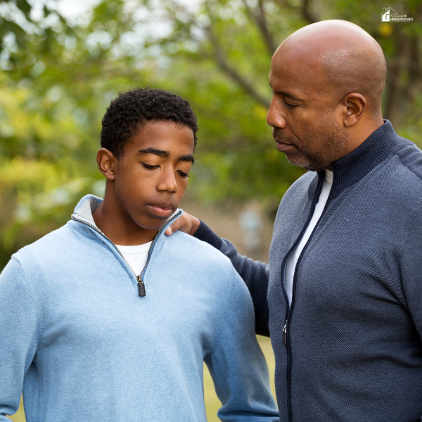 A father gently speaks to his son outdoors, placing a comforting hand on his shoulder during a serious moment.