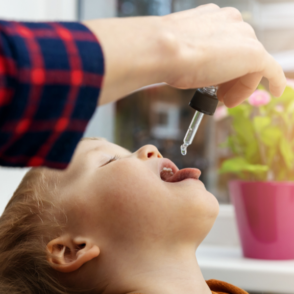A child opens their mouth as an adult administers allergy drops under the tongue using a small dropper.
