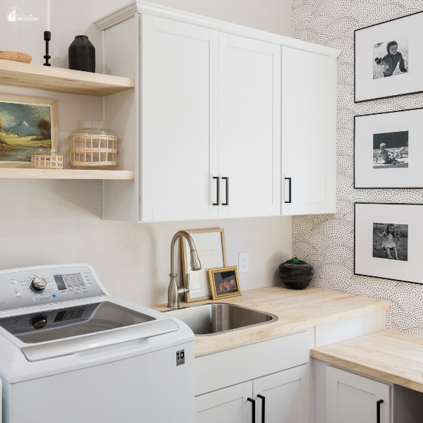 A bright laundry room featuring white cabinetry, wooden countertops, a stainless steel sink, and decorative wall art.