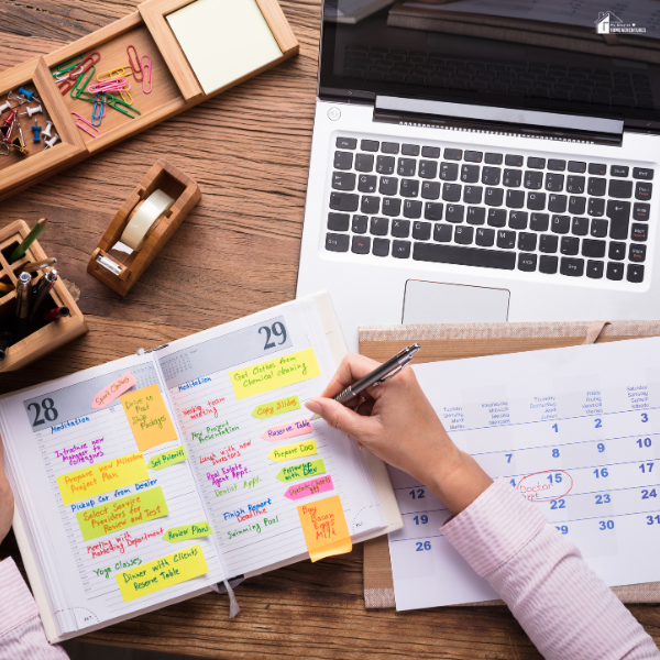 A person writes on a calendar while sitting at a desk with a laptop, planner, and colorful sticky notes.