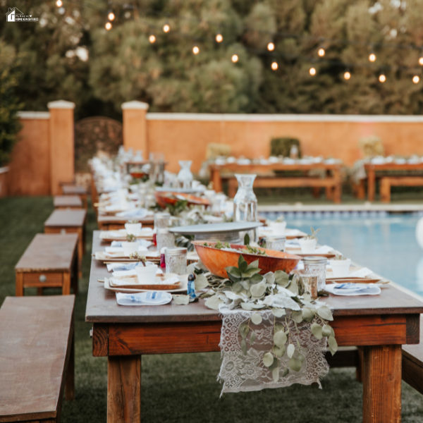 Long decorated tables set up for a backyard wedding reception beside a pool with string lights creating a cozy ambiance.