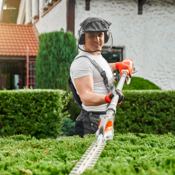 A man uses a long-reach hedge trimmer to cut tall hedges in a residential garden.