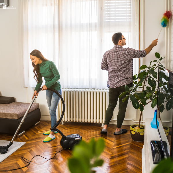 Couple cleaning a bright living room together in a tenancy setup, with one vacuuming the floor and the other dusting near a window.