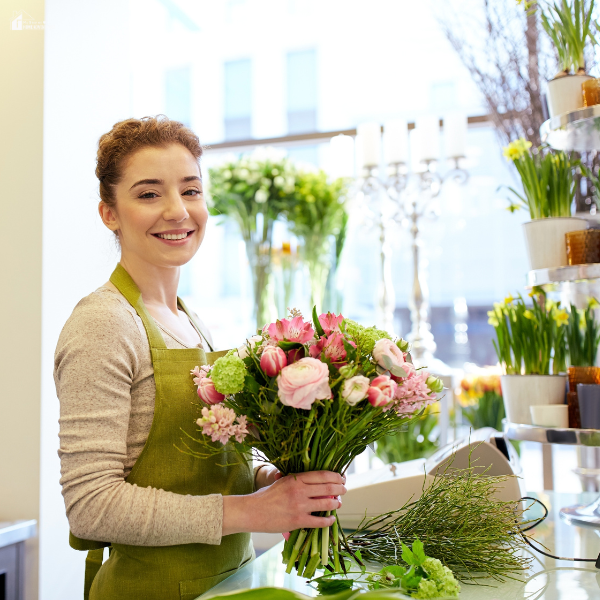 A smiling florist in a green apron holding a bouquet of pink and white flowers inside a flower shop.