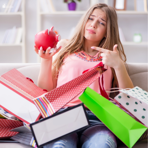 A young woman surrounded by colorful shopping bags holding a piggy bank and pointing at it with a concerned expression.