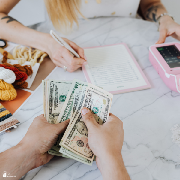 Person counting a stack of cash while another writes on a notepad at a table with craft supplies.