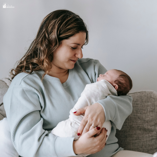 A young mother lovingly holds and looks down at her newborn baby while sitting on a couch.