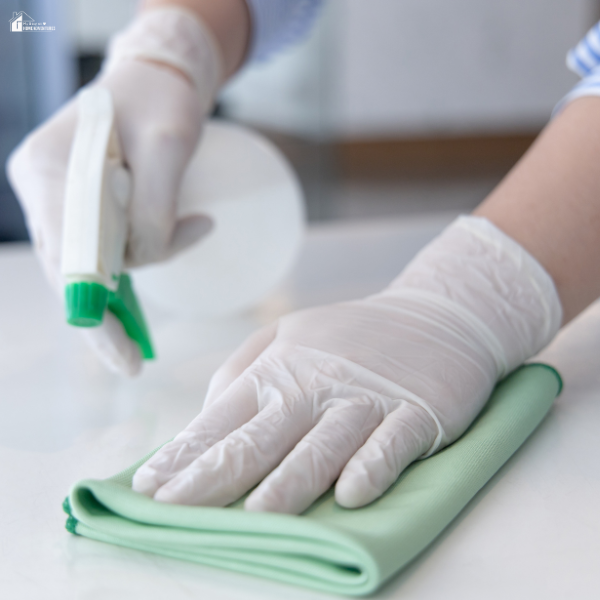Person wearing gloves using a spray bottle and microfiber cloth to clean a white surface.