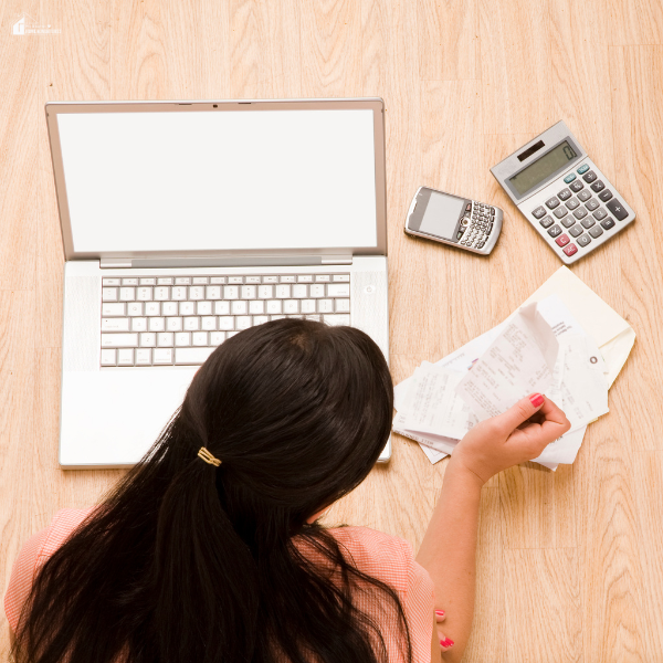 A woman sitting on the floor in front of a laptop, holding receipts with a calculator and phone nearby, reviewing her expenses.