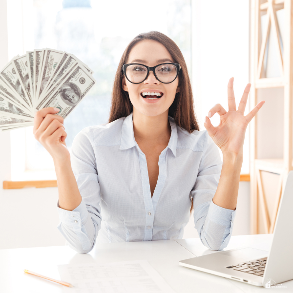 A smiling woman sitting at a desk holding a fan of cash in one hand and making an OK sign with the other, representing financial success.