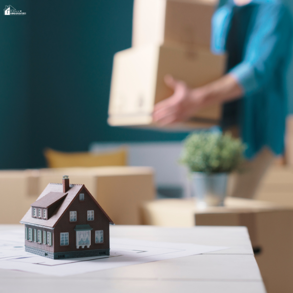 A miniature house model sits on a table with moving boxes and a person carrying a box in the background, symbolizing relocation.