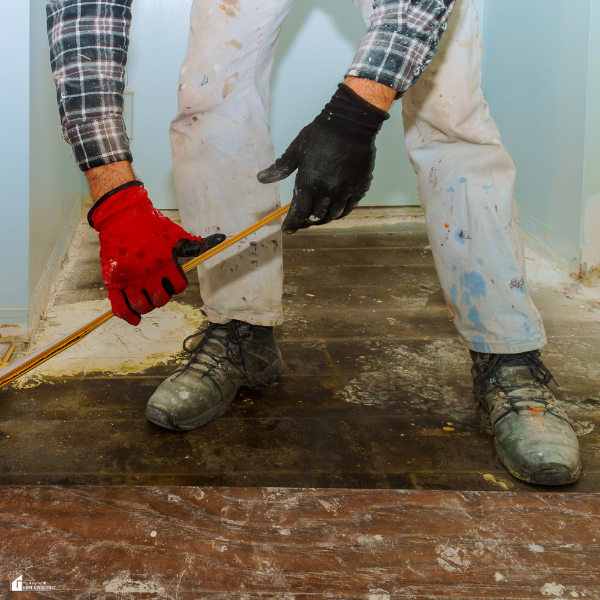 A worker wearing gloves and protective clothing removes old flooring with a tool during a renovation.