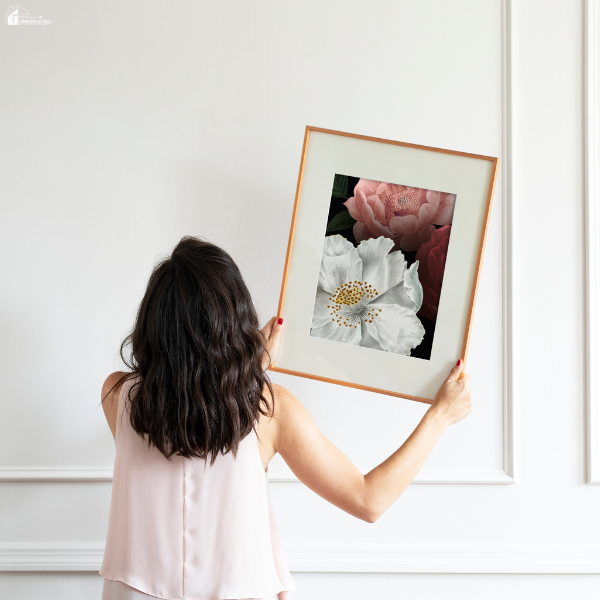 A woman holding up a framed floral print against a white wall, preparing to hang seasonal wall art.