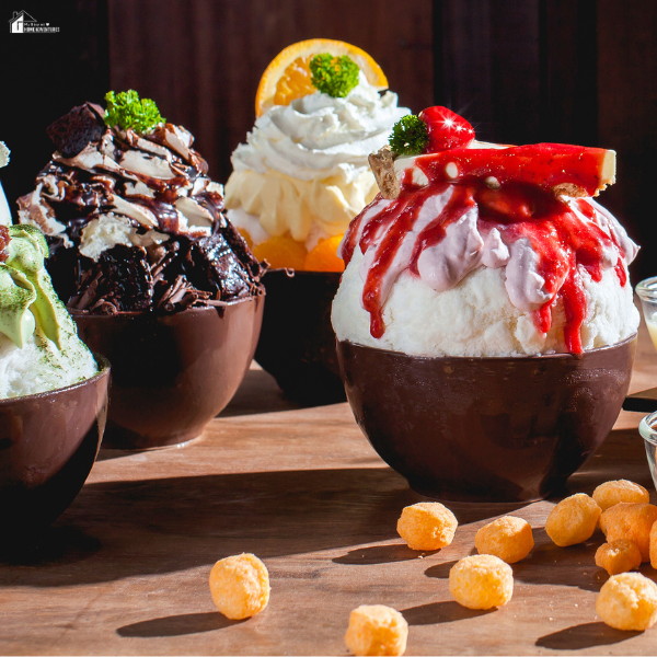 A variety of snowflake ice cream bowls topped with whipped cream, chocolate, and fruit served on a wooden table.