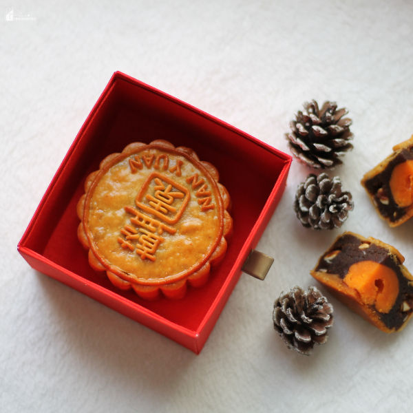 A traditional mooncake in a red box with pinecones and sliced mooncake pieces beside it.