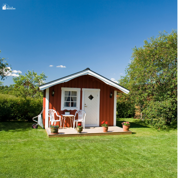 A small red garden room with white trim, a front porch, and lawn chairs, set in a lush green backyard under a clear blue sky.