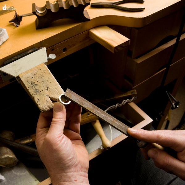 A person using a flat file to shape a small metal ring at a jeweler’s bench with various tools nearby.
