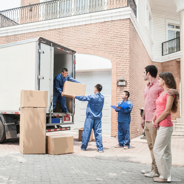 Professional movers in blue uniforms load boxes into a moving truck as a couple watches outside a suburban home.