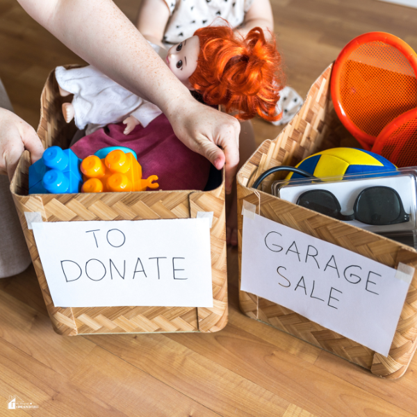 Close-up of hands sorting children’s toys and household items into labeled donation and garage sale baskets on a wooden floor.