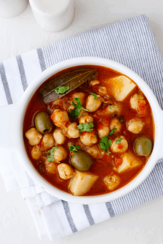 A white bowl filled with garbanzos guisados, featuring chickpeas, olives, potatoes, and bay leaf on a striped cloth.