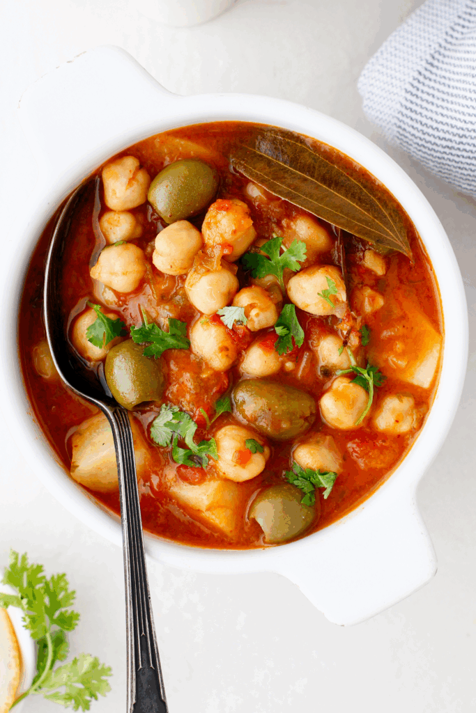 Overhead view of Puerto Rican chickpea stew with a spoon in a white bowl, showing hearty vegetables and fresh herbs.