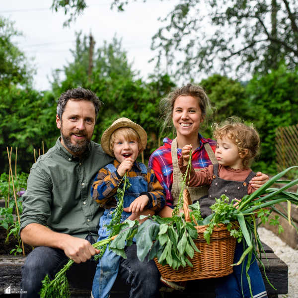 Family of four holding fresh vegetables and sitting in a home garden filled with greenery.