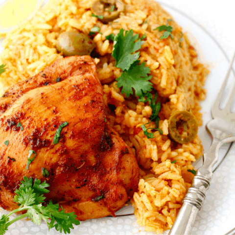 A serving of arroz con pollo on a white plate with golden rice, green olives, tender chicken thigh, fresh cilantro, and lime wedge.