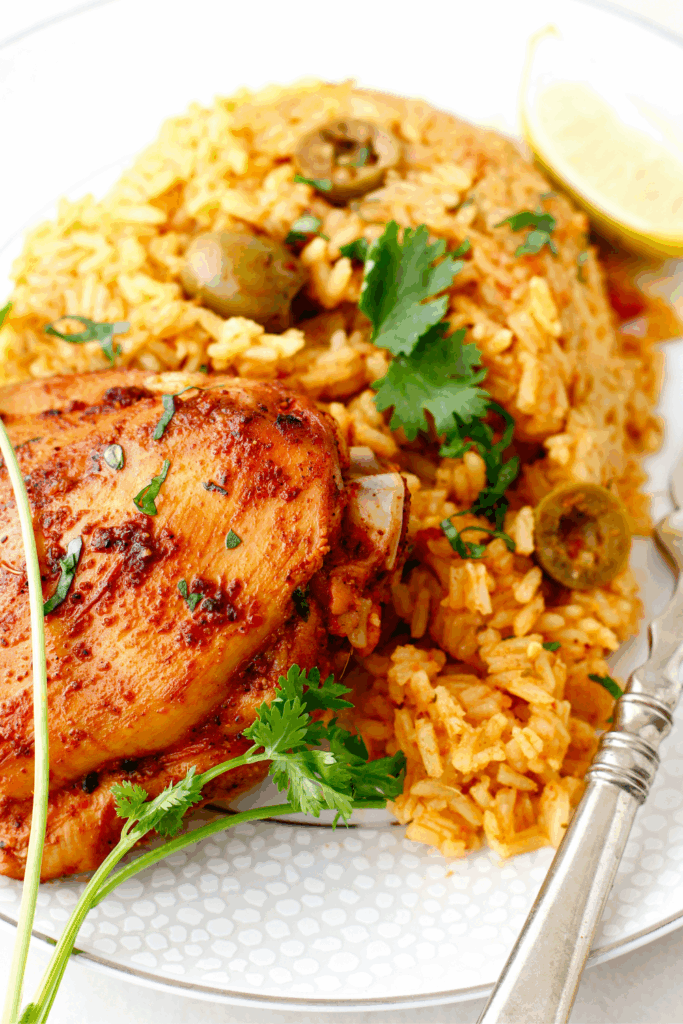 A serving of arroz con pollo on a white plate with golden rice, green olives, tender chicken thigh, fresh cilantro, and lime wedge.