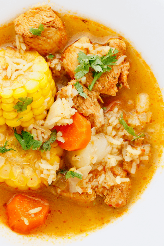 Close-up of plated Puerto Rican chicken soup with rice, corn, carrots, and lemon wedge in a white bowl.