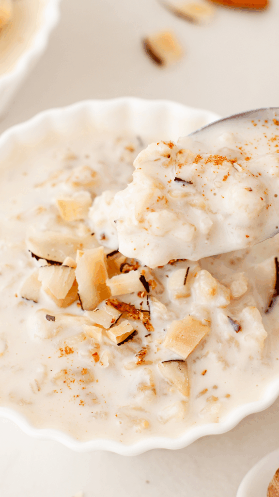 A close-up of a spoonful of coquito oatmeal lifted from a white bowl, showing its thick, creamy texture and coconut topping.