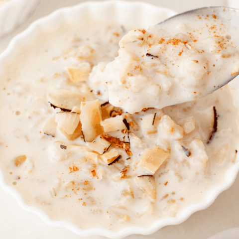 A close-up of a spoonful of coquito oatmeal lifted from a white bowl, showing its thick, creamy texture and coconut topping.