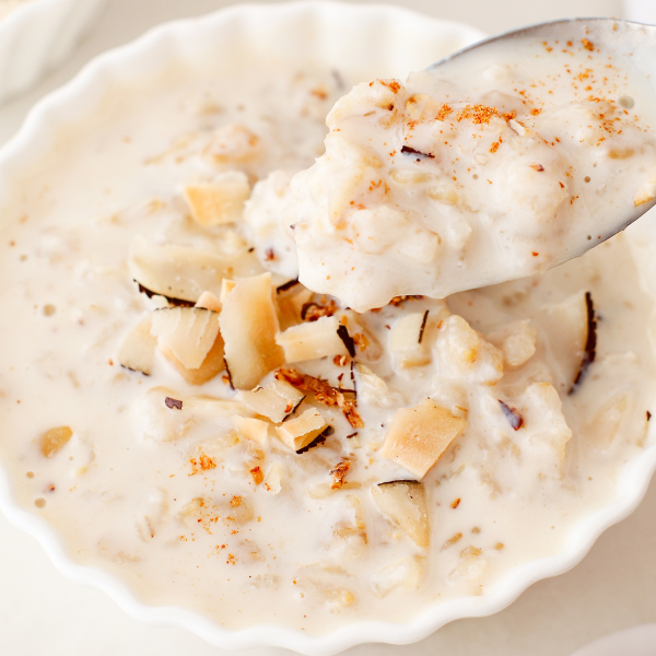 A close-up of a spoonful of coquito oatmeal lifted from a white bowl, showing its thick, creamy texture and coconut topping.