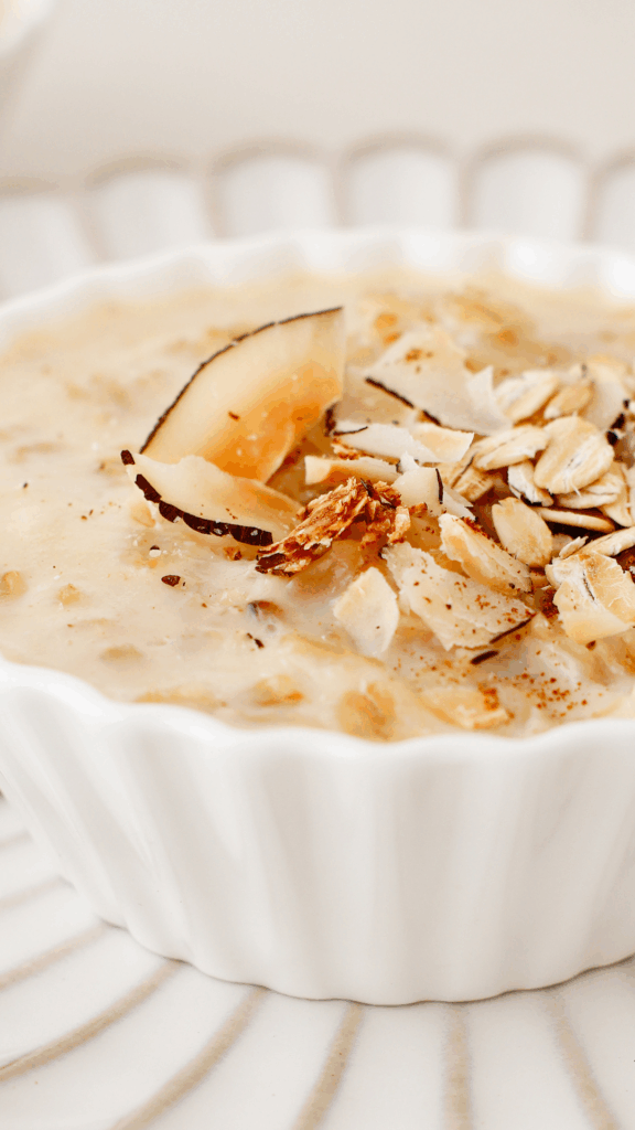 A white bowl of coquito oatmeal garnished with toasted coconut slices and oats, placed on a striped plate.