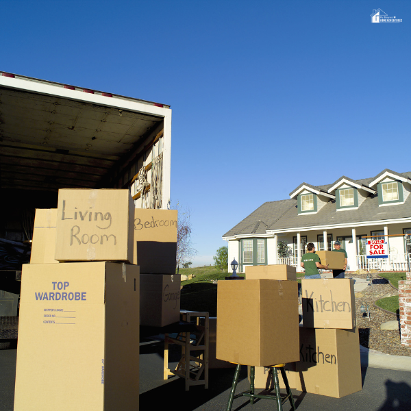 Labeled cardboard boxes are stacked near a moving truck outside a suburban home.