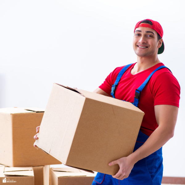 A smiling mover in a red uniform carries a cardboard box in a brightly lit room.