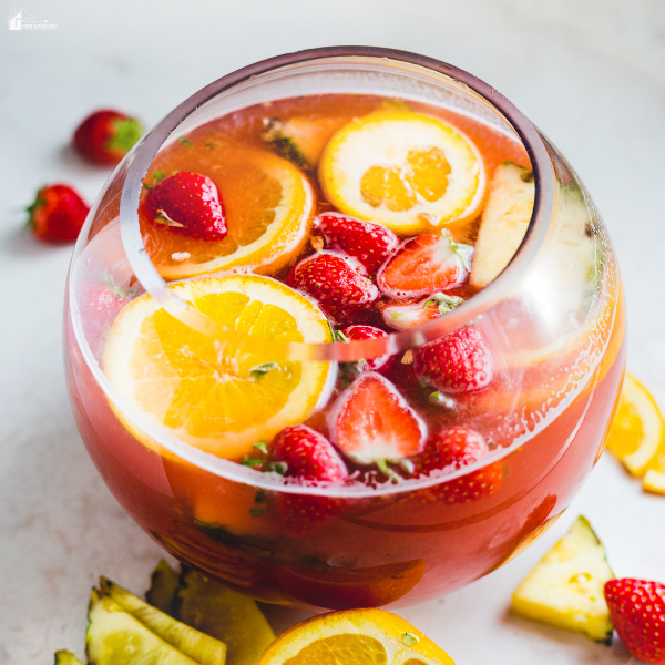 A large glass bowl filled with a colorful fruit punch made with sliced oranges, strawberries, and pineapple chunks.