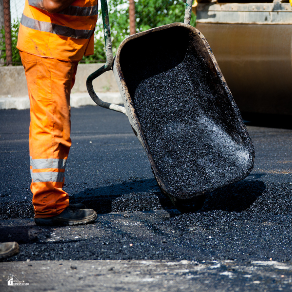 A construction worker pours fresh asphalt from a wheelbarrow onto a road surface during paving work.