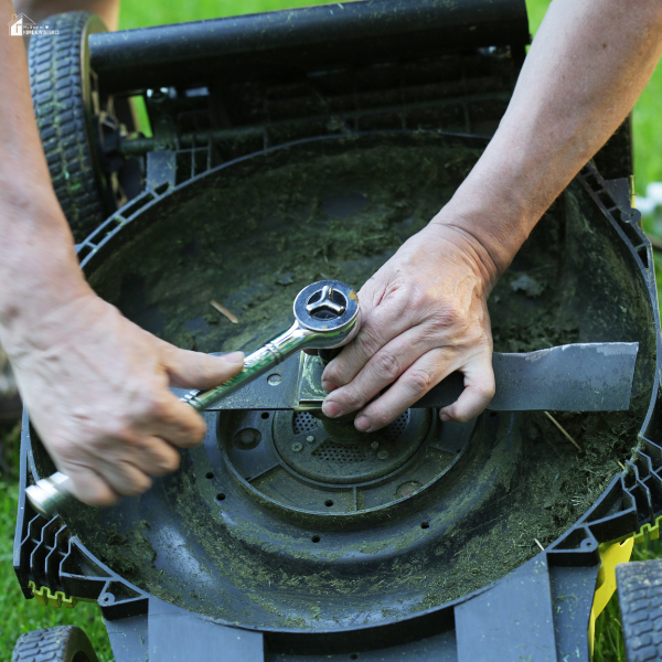 Close-up of hands using a wrench to adjust or replace the blade on the underside of a lawn mower.